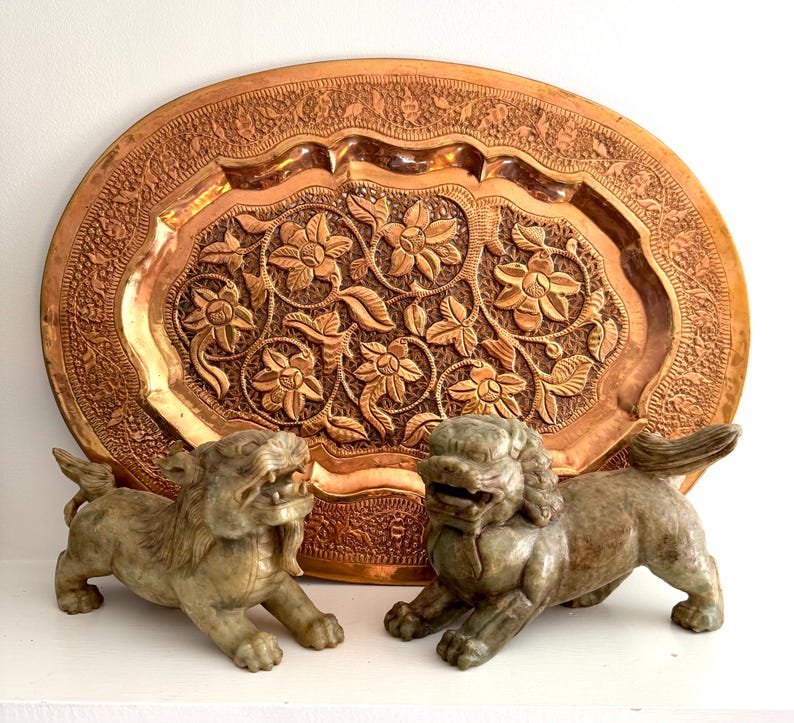 May include: A large, oval copper tray with intricate floral carvings. Two stone Foo Dog statues are in front of the tray. The tray and statues are on a white surface.