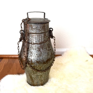 May include: A large, antique, silver-colored metal container with a lid and a chain handle. The container has a textured surface and is sitting on a white fluffy rug.