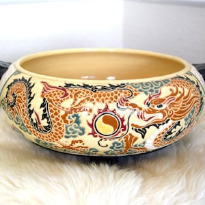 Puede incluir: Un cuenco grande de cerámica de color crema con dos asas de dragón negro. El cuenco está decorado con dos dragones, nubes y un símbolo yin-yang en tonos marrones, rojos, azules y negros.