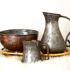 May include: A set of three antique copper and brass serving pieces, including a large bowl, a pitcher, and a mug. The pieces are all decorated with intricate designs and have a worn, aged patina.