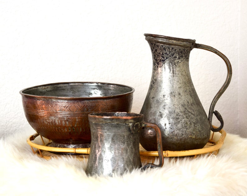 May include: A set of three antique copper and brass serving pieces, including a large bowl, a pitcher, and a mug. The pieces are all decorated with intricate designs and have a worn, aged patina.