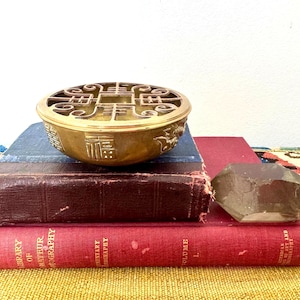 May include: A round brass incense burner featuring an intricate openwork lid with geometric patterns and embossed Chinese characters on the side. The bowl has a polished metallic finish and sits atop a stack of two vintage hardcover books.