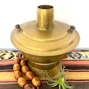 May include: A vintage brass samovar featuring a wide, rounded body with a central chimney, a removable lid with two small dark wooden knobs, and a perforated base. The metallic surface shows a warm, aged patina with visible signs of wear.