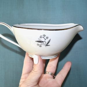 May include: White ceramic gravy boat with a black and gray floral design. The gravy boat has a gold trim around the rim and handle. The handle is curved and the spout is pointed.