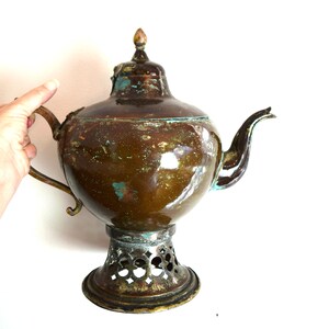 May include: A large, antique copper teapot with a decorative stand. The teapot has a rounded body and a long spout. The stand is made of metal and has an intricate design.