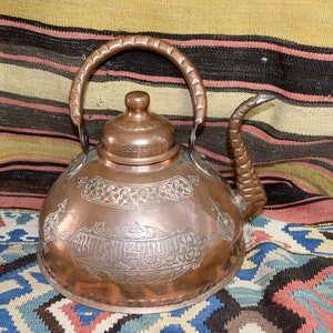 May include: A copper teapot with an ornate design and a long spout. The teapot is decorated with intricate patterns and Arabic script.