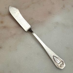 May include: A silver-toned metal butter knife featuring a flat, wide blade and a slender handle. The handle is decorated with a textured, hammered finish and an engraved shield-shaped emblem containing the letter B, surrounded by ornate detailing.