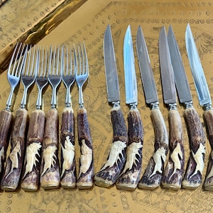 May include: A collection of vintage silverware, including forks and knives, with ornate handles. The handles are a dark brown color with carved white accents. The blades are silver and the set is arranged on a gold-toned tray.