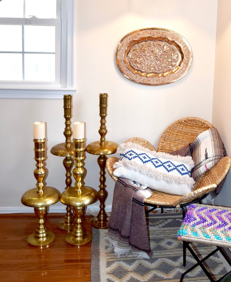 May include: A cozy interior scene featuring gold candle holders, a woven chair with a patterned pillow and blanket, and a decorative copper tray on the wall. A patterned rug and small stool complete the bohemian aesthetic.