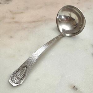 May include: A silver-toned metal ladle featuring a hammered texture along the handle. The handle terminates in a decorative shield-shaped emblem with engraved floral details and the letters "DED" centered within the shield design.