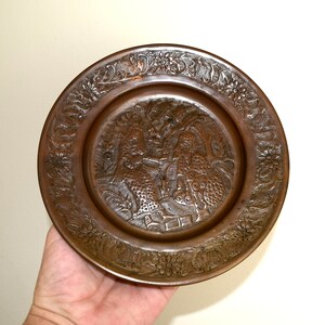 May include: A round copper plate with an intricate design of two figures in a garden setting. The plate has a raised rim and a smooth, polished surface.