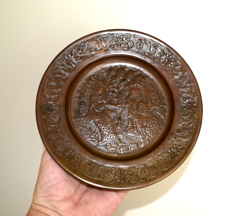 May include: A round copper plate with an intricate design of two figures in a garden setting. The plate has a raised rim and a smooth, polished surface.