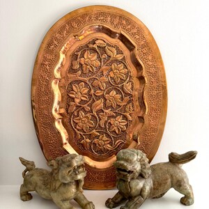 May include: A large, oval copper tray with intricate floral and leaf carvings. Two stone lion statues are in front of the tray. The tray and statues are on a white surface.