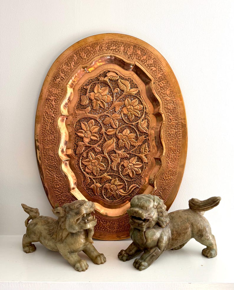 May include: A large, oval copper tray with intricate floral and leaf carvings. Two stone lion statues are in front of the tray. The tray and statues are on a white surface.
