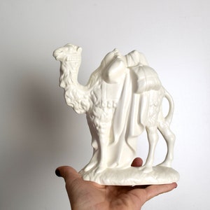 May include: A white ceramic camel figurine with a saddle and bags. The camel is standing on a base and has a detailed head and body.