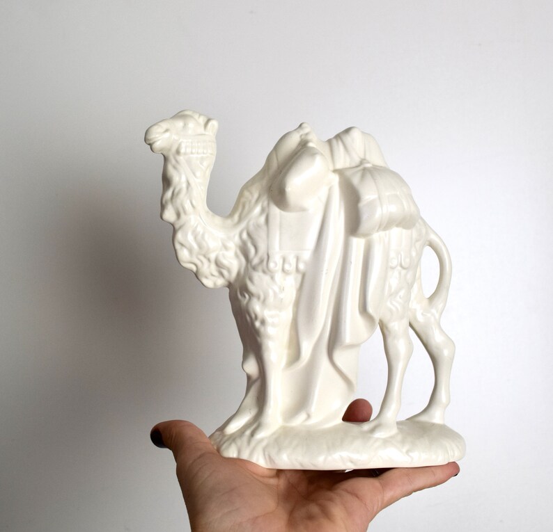 May include: A white ceramic camel figurine with a saddle and bags. The camel is standing on a base and has a detailed head and body.