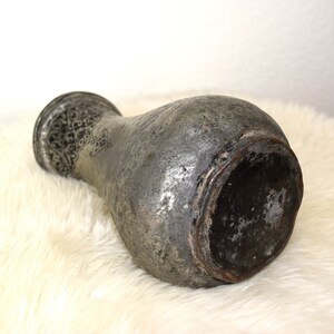 May include: A silver metal vase with intricate engraved designs. The vase has a rounded body and a wide mouth. The vase is sitting on a white fluffy surface.