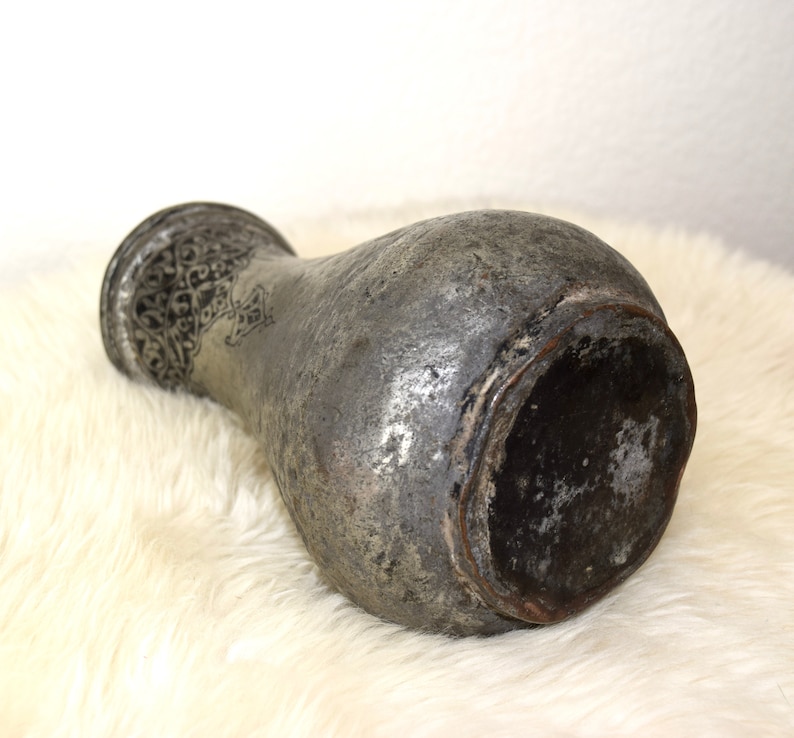 May include: A silver metal vase with intricate engraved designs. The vase has a rounded body and a wide mouth. The vase is sitting on a white fluffy surface.