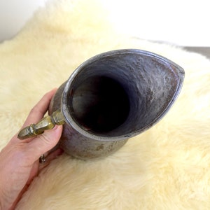 May include: A vintage pewter pitcher with a hammered finish and a brass handle. The pitcher has a wide mouth and a rounded body.