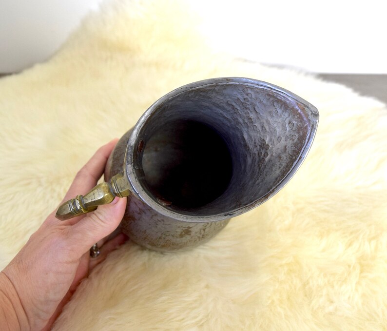 May include: A vintage pewter pitcher with a hammered finish and a brass handle. The pitcher has a wide mouth and a rounded body.