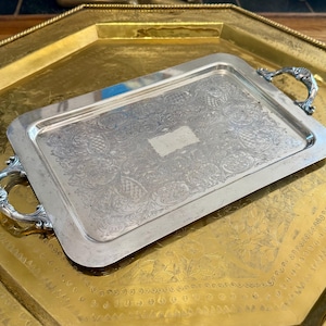 May include: A rectangular silver-toned serving tray featuring ornate engraved scrollwork patterns across the surface, a smooth central rectangular area, and two decorative metal handles with shell-like detailing on each of the shorter sides.