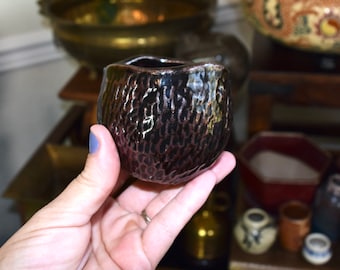 Puede incluir: Una pequeña maceta de cerámica marrón oscuro con una superficie texturizada. La maceta tiene una forma redondeada y un borde ligeramente acampanado.