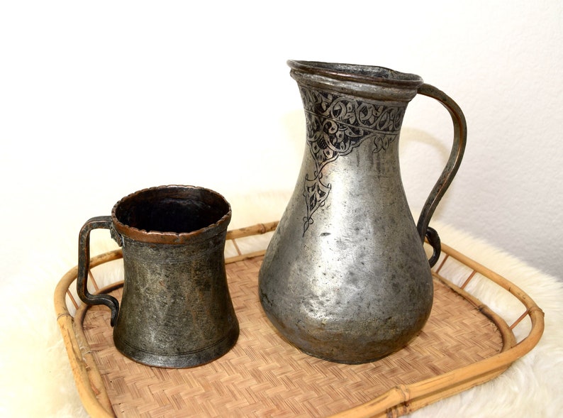 May include: A set of two antique copper pitchers with intricate etched designs. The larger pitcher has a wide mouth and a handle, while the smaller pitcher is a mug with a single handle. Both pitchers are on a woven bamboo tray.