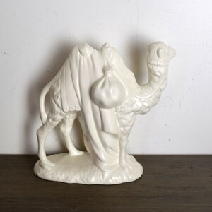 May include: White ceramic camel figurine with a saddle and a bag on its back. The camel is standing on a base with a textured surface.