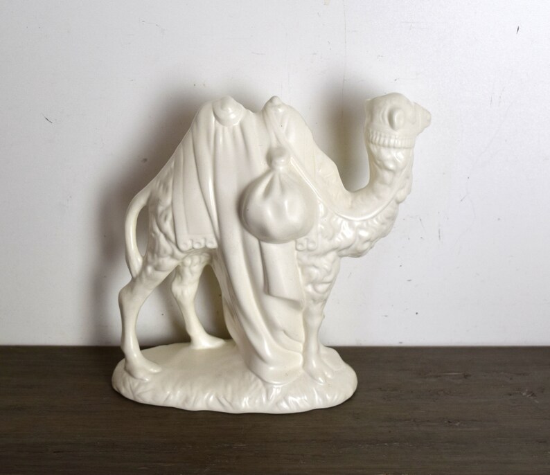May include: White ceramic camel figurine with a saddle and a bag on its back. The camel is standing on a base with a textured surface.