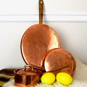 Op de afbeelding: Een set van drie koperen potten met messing handvatten. De grootste pot heeft een brede, platte bodem en een afgeronde bovenkant. De twee kleinere potten hebben een traditionelere vorm. De potten staan op een wit, pluche oppervlak met twee citroenen.