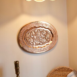 May include: A copper-colored, oval-shaped wall hanging with an intricate floral design. The decorative piece is mounted on a neutral-colored wall. The design features raised floral patterns within a detailed border. The piece is likely made of metal.