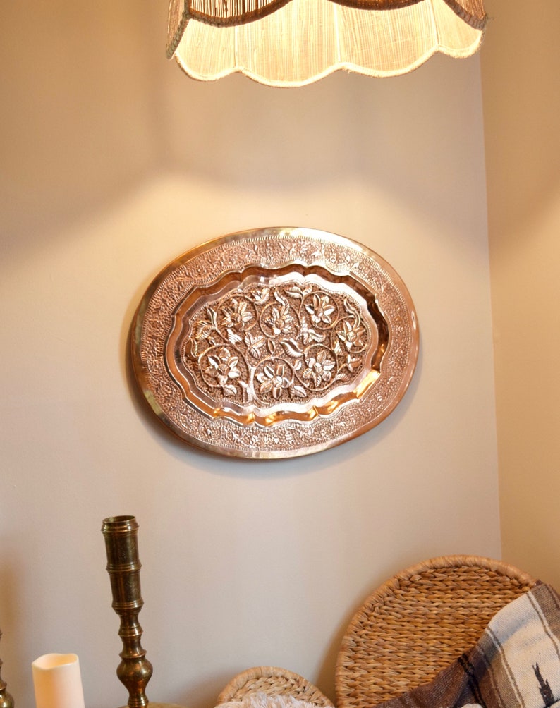 May include: A copper-colored, oval-shaped wall hanging with an intricate floral design. The decorative piece is mounted on a neutral-colored wall. The design features raised floral patterns within a detailed border. The piece is likely made of metal.