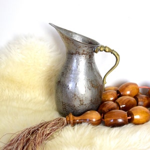 May include: A silver pitcher with a brass handle sits on a white fluffy rug with a wooden beaded garland.