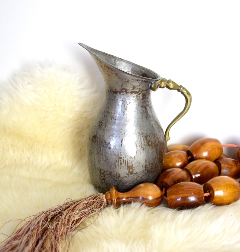 May include: A silver pitcher with a brass handle sits on a white fluffy rug with a wooden beaded garland.