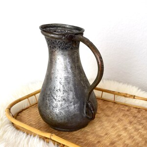 May include: A silver pitcher with an ornate design around the top and a copper handle. The pitcher is sitting on a woven bamboo tray.