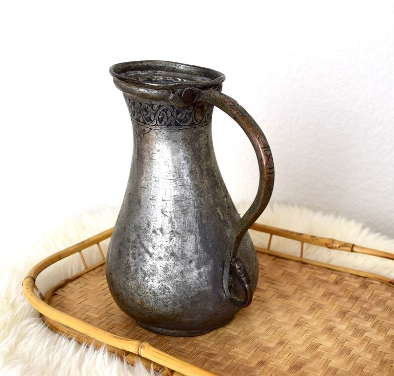 May include: A silver pitcher with an ornate design around the top and a copper handle. The pitcher is sitting on a woven bamboo tray.