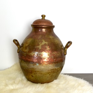 May include: A large, antique copper and brass pot with two handles and a lid. The pot has a hammered finish and is slightly tarnished.
