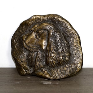 May include: A bronze sculpture of a dog's head, the dog is facing left and has long, flowing ears. The sculpture is mounted on a rough, textured base.