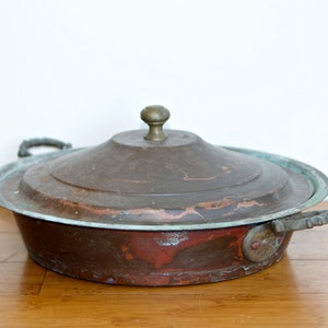 May include: A vintage copper pot with a lid and two handles. The pot has a patina and is slightly tarnished. The lid has a brass knob.