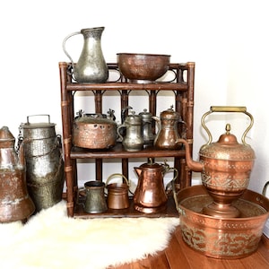May include: A collection of antique copper and silver teapots, pitchers, and bowls arranged on a wooden shelf. The items are all in various sizes and shapes, and some have intricate designs.