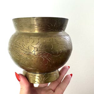 May include: A round brass bowl featuring an etched design of a bird in flight surrounded by swirling floral patterns. The metal has a muted gold tone with a wide opening and a flared base, showcasing detailed engravings on the exterior surface.