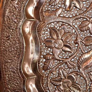 May include: Close-up of a copper-colored decorative tray with intricate floral and leaf patterns. The tray features a raised, wavy border and detailed openwork designs, showcasing a vintage aesthetic.
