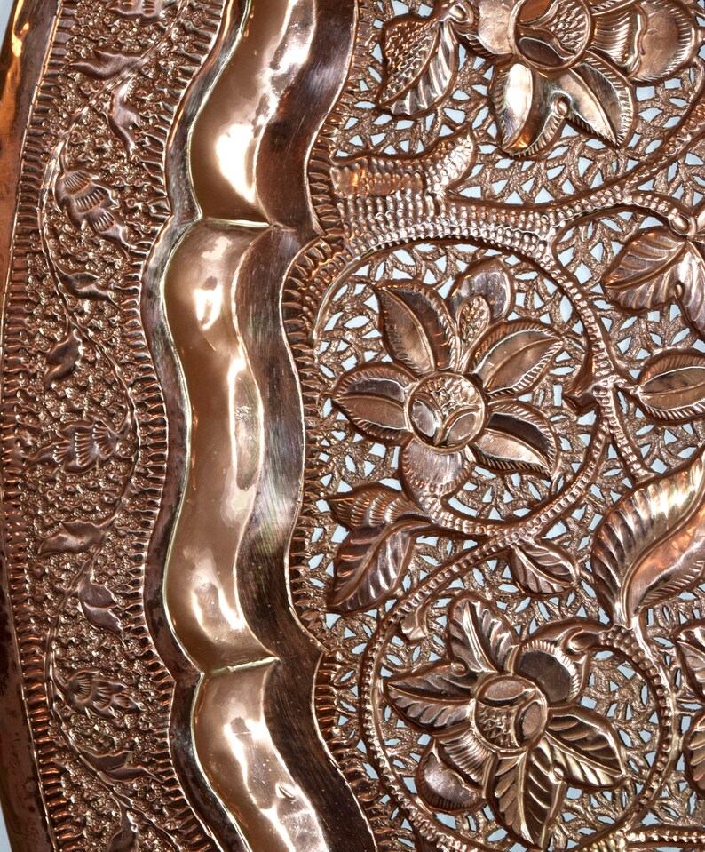May include: Close-up of a copper-colored decorative tray with intricate floral and leaf patterns. The tray features a raised, wavy border and detailed openwork designs, showcasing a vintage aesthetic.