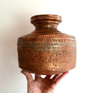 Può includere: Un vaso di rame con intricati motivi floreali e geometrici incisi sulla superficie. Il vaso ha un corpo arrotondato e una bocca larga.