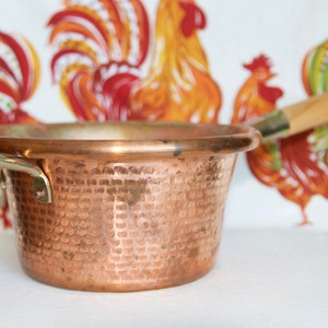 May include: A hammered copper saucepan with a brass handle. The saucepan has a textured surface and is sitting on a white surface.