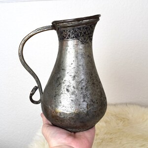 May include: A silver pitcher with a decorative design around the top. The pitcher has a handle and a rounded body.