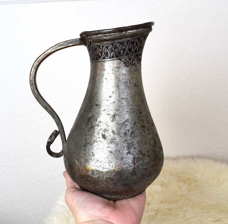 May include: A silver pitcher with a decorative design around the top. The pitcher has a handle and a rounded body.