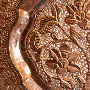 May include: Close-up of a copper-colored decorative plate with an intricate floral and leaf design. The plate features embossed flowers and a detailed openwork pattern, creating a textured surface. The plate has a curved edge.