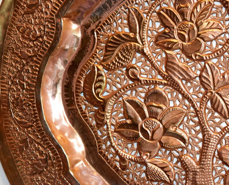 May include: Close-up of a copper-colored decorative plate with an intricate floral and leaf design. The plate features embossed flowers and a detailed openwork pattern, creating a textured surface. The plate has a curved edge.
