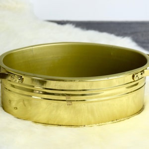 Puede incluir: Una tina de metal ovalada dorada con dos asas. La tina está vacía y tiene un acabado brillante.